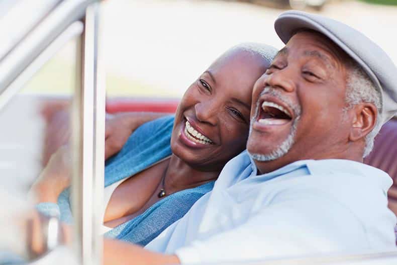 A 55+ couple laughing an smiling while driving in the car on a road trip.