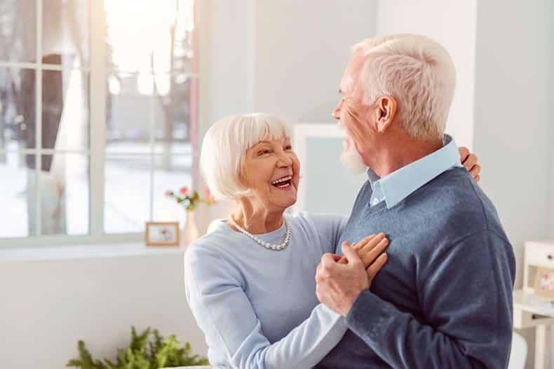 A joyful elderly husband and wife dancing in the living room while smiling.