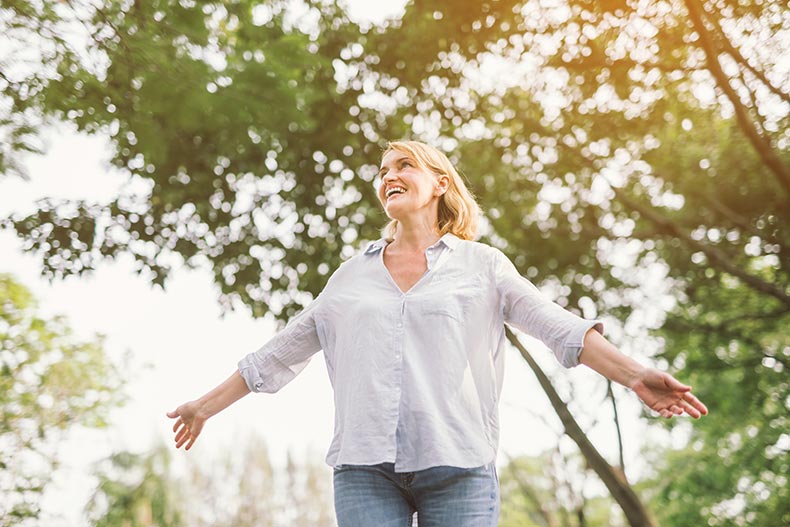 A happy 55+ woman in a spring or summer forest park with open arms.