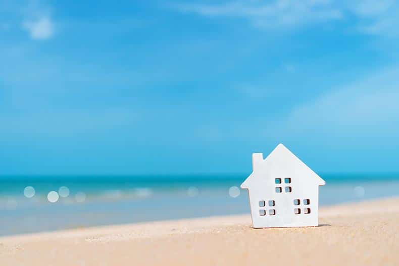 Closed up of a tiny home model on sand with sunlight and a beach in the background.