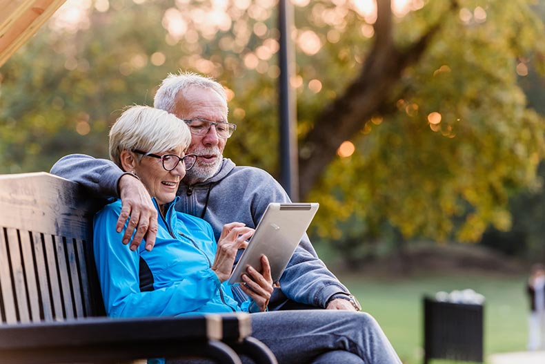 A senior couple sitting on a bench and reading 55+ community reviews on a tablet.