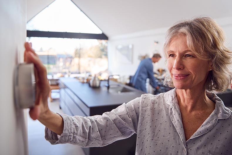 A 55+ woman adjusting a wall mounted digital thermostat at home.