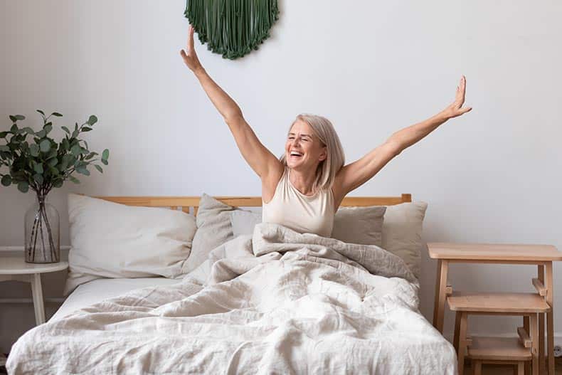 55+ woman wakes up and stretches in bed in light bedroom at home.