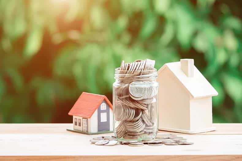 A jar of coins beside two house models with a green background.