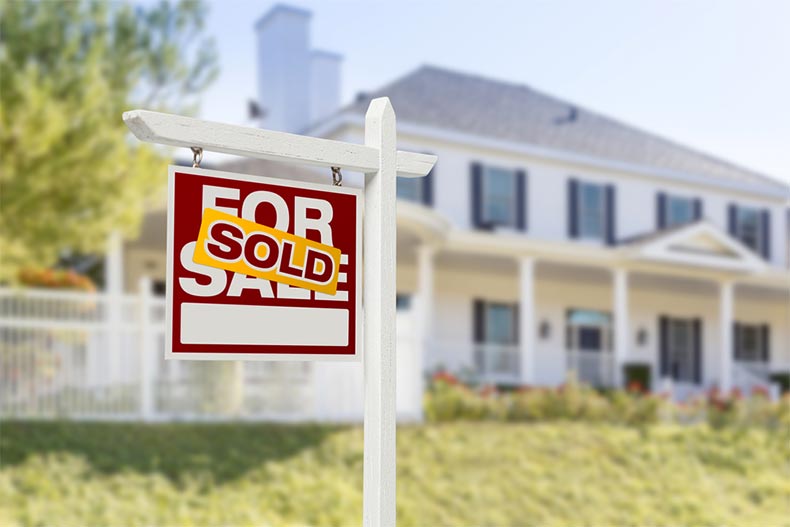 A SOLD sticker over a "For Sale" sign in front of a house.