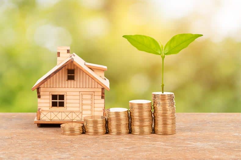 A plant growing out of a stack of coins beside a wooden model house.