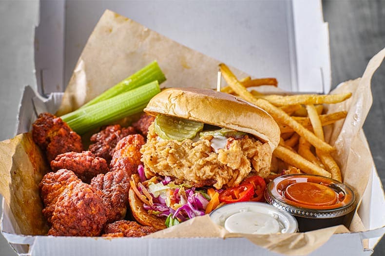 A take out box with a fried chicken sandwich, boneless wings, and french fries.