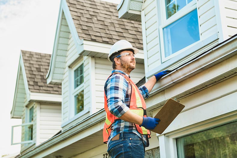 A man with hard hat standing on steps inspecting house roof.