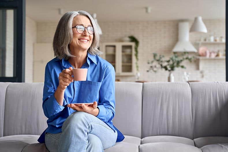 A smiling woman relaxing with a cup of tea in her 55+ apartment.