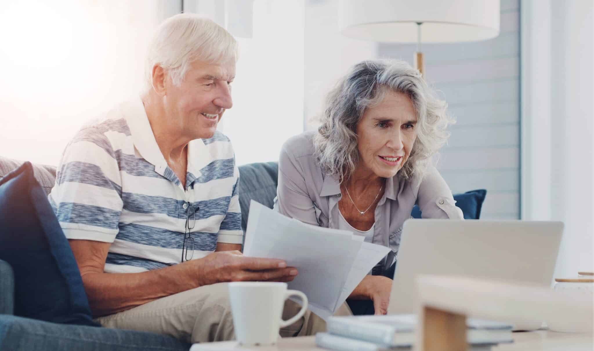 Older couple holding papers and leaning over to look at a laptop.