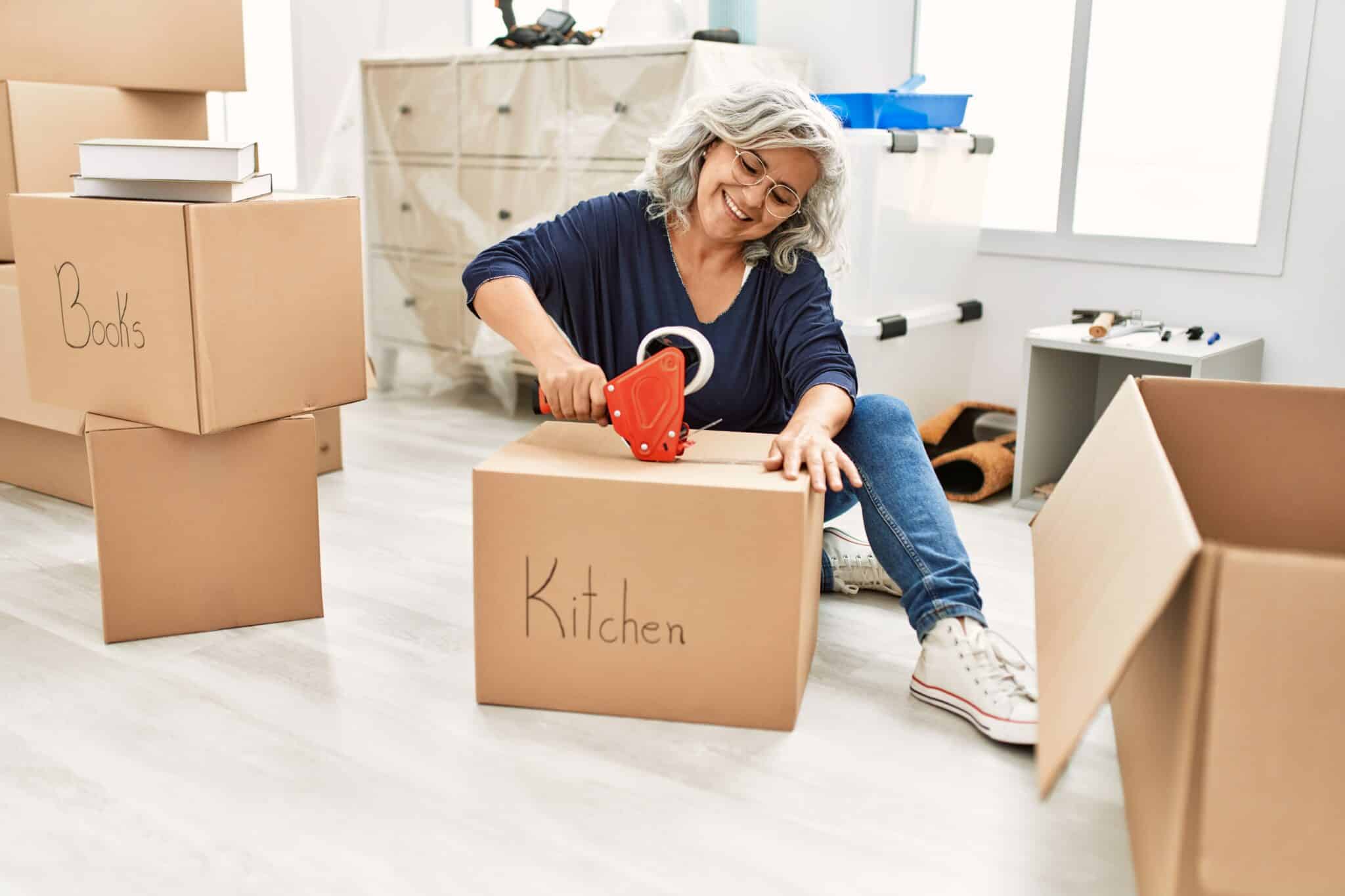 A 55+ woman smiling while packing a kitchen cardboard box in a new home.