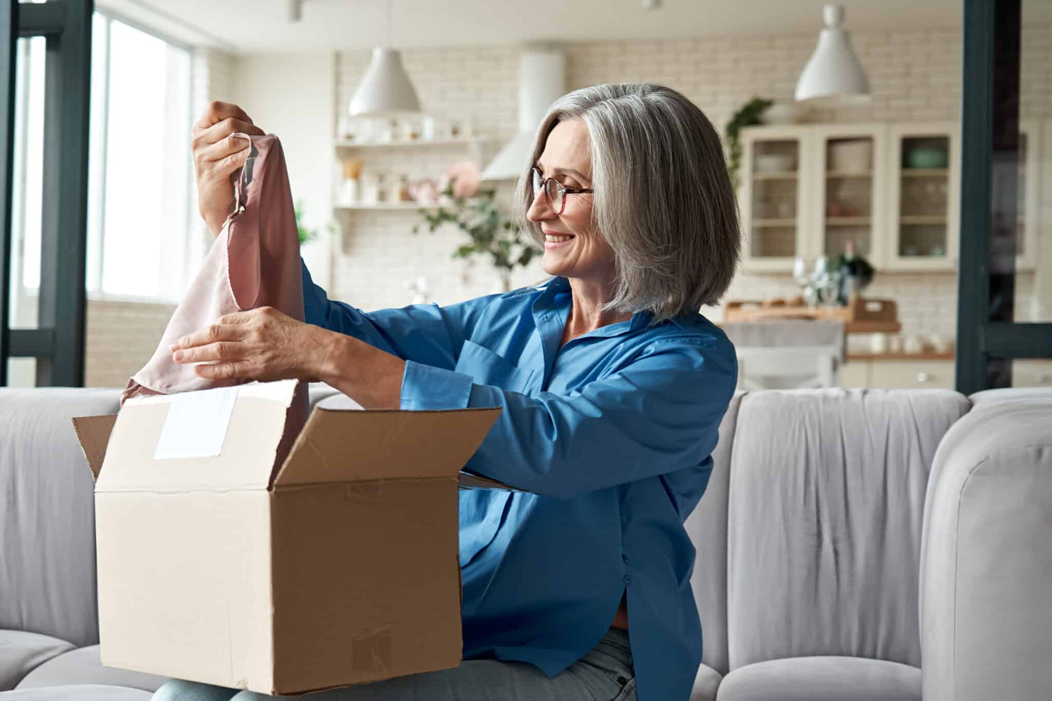Woman with gray hair and glasses unpacking a cardboard box on the couch.