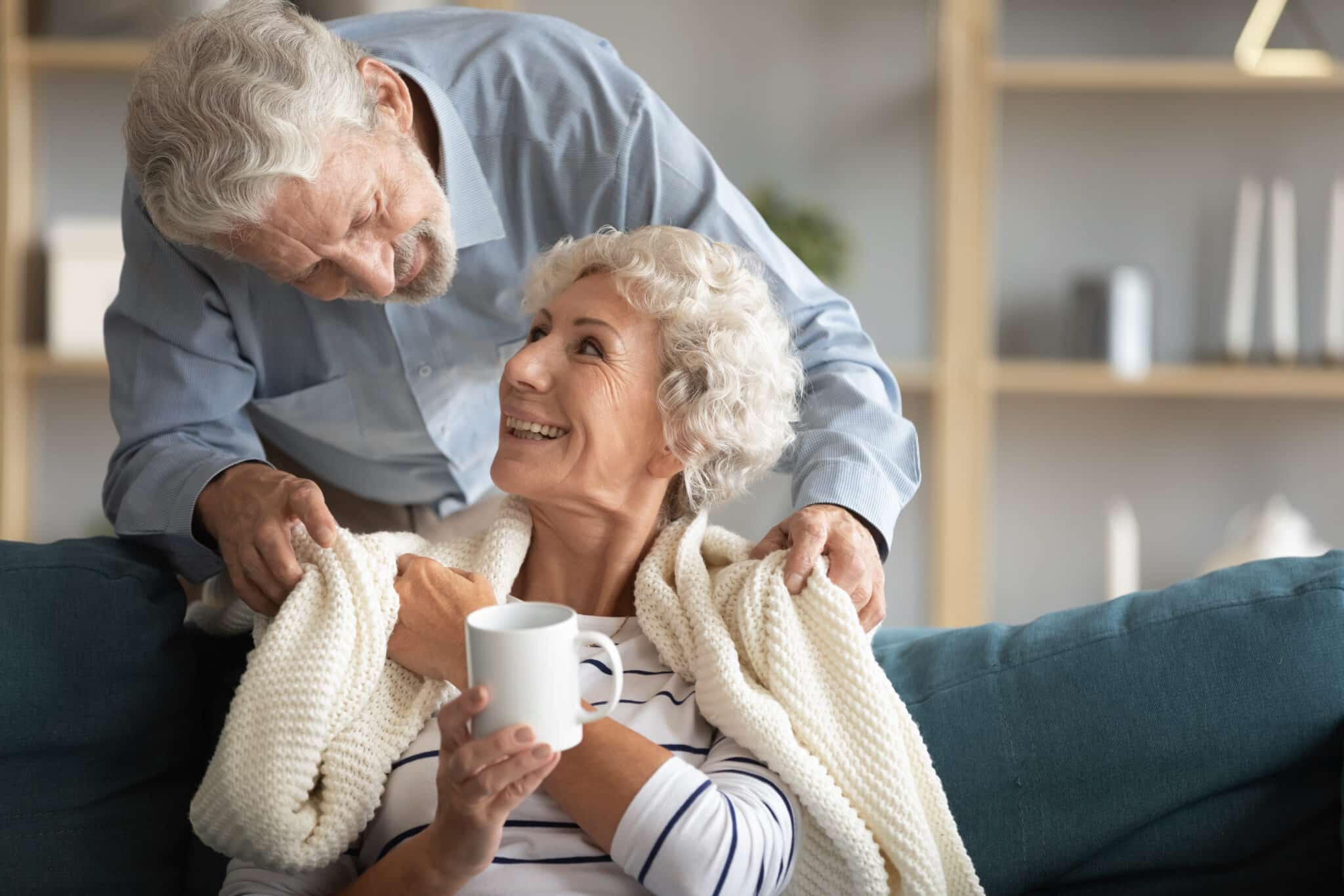 Senior man covering the shoulders of a senior woman with a blanket on the couch.