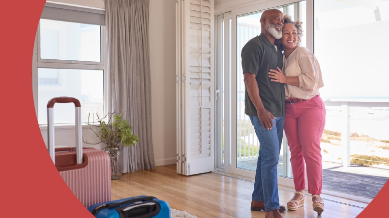 A 55+ couple with luggage arriving at a beachfront house overlooking the ocean.