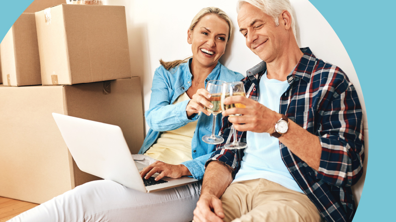 A happy mature couple using a laptop and drinking wine together on moving day.