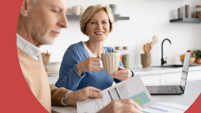 A happy retired woman smiling near her husband while he looks over their Social Security Benefits.