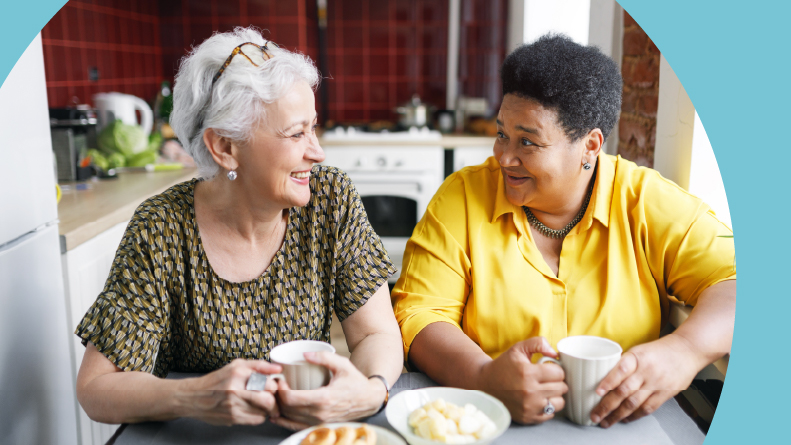 Two 55+ ladies sitting at a kitchen table and drinking coffee.