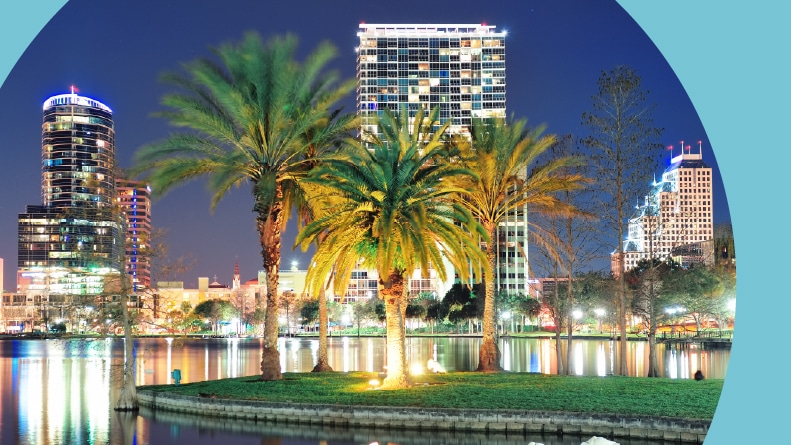 View of Downtown Orlando and Lake Eola at night with skyscrapers, palm trees, and a clear sky.