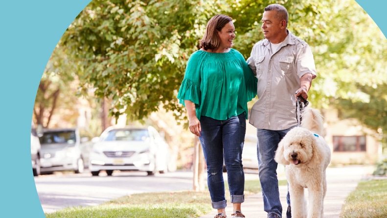 A senior couple walking their dog along a suburban sidewalk on a sunny day.
