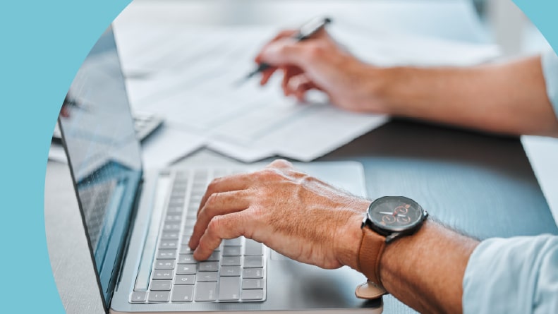 A desk, a laptop, and hands with paperwork in an office doing taxes.