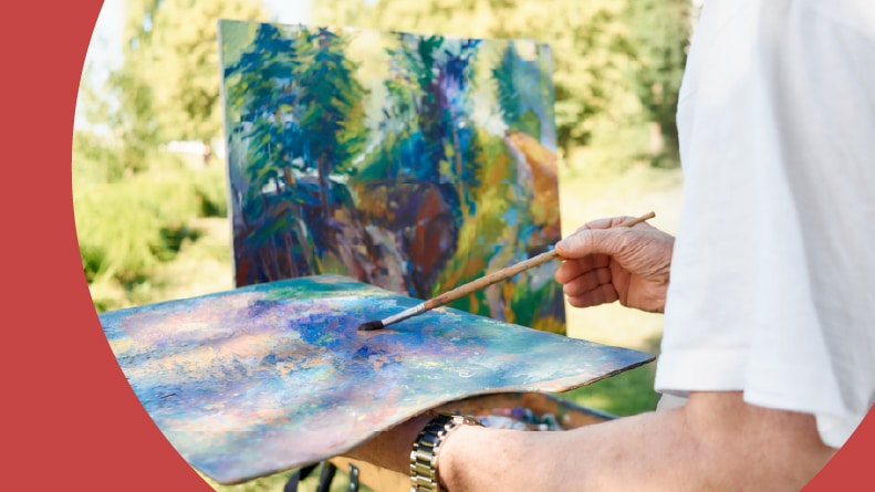 Closeup of a senior man's hands holding a painting brush and a palette while painting in a park.