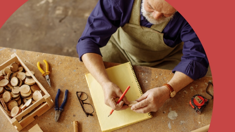 Top view of a senior man taking measurements with a pencil and notebook while woodworking.