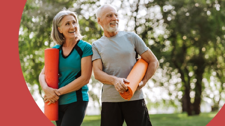 A 55+ couple holding yoga mats and smiling in the park.