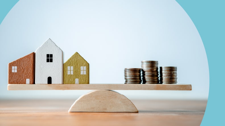 A balance scale made of a wooden plank with small wood homes and a stack of coins.