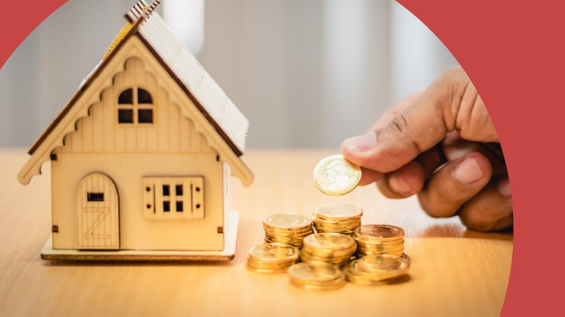 A man's hand stacking gold coins next to a wood model house.