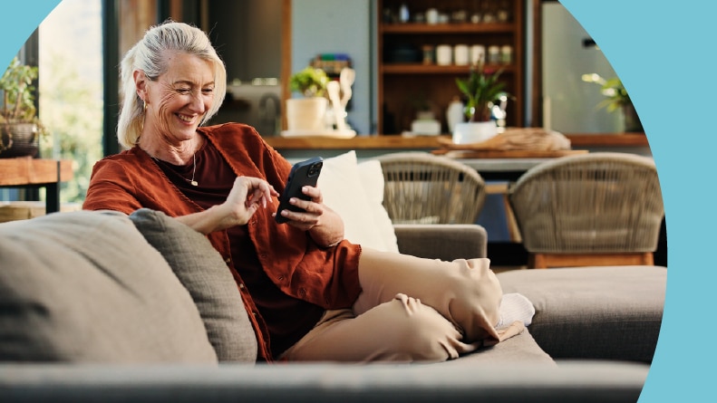 A happy 55+ woman reviewing her finances on her phone while sitting on the couch at home.