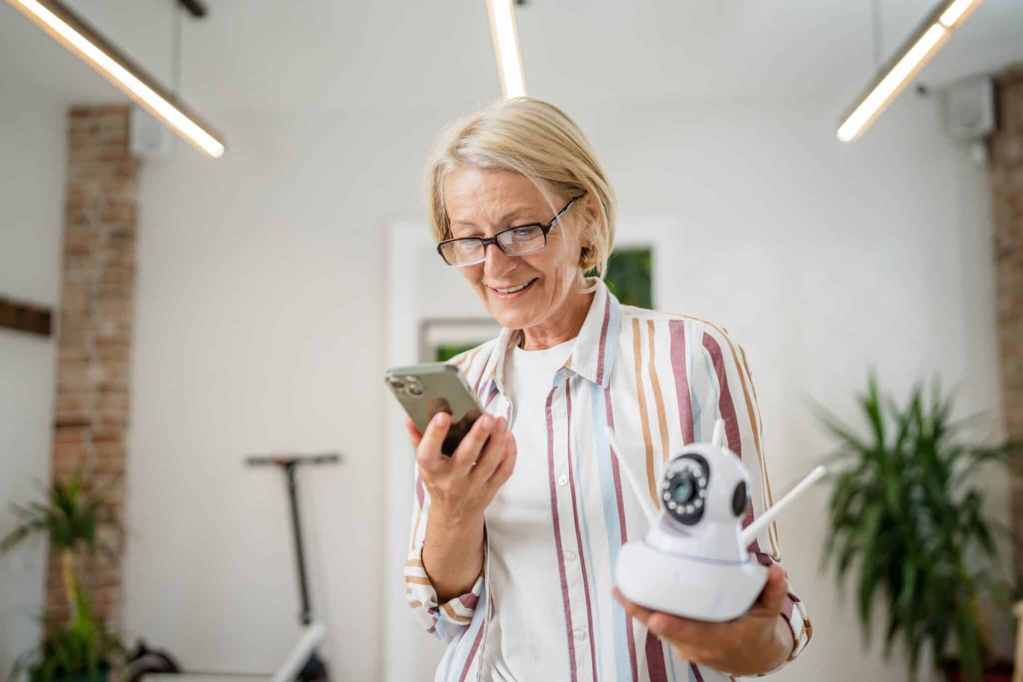 A 55+ woman holding a modern security surveillance camera connected to her mobile phone.