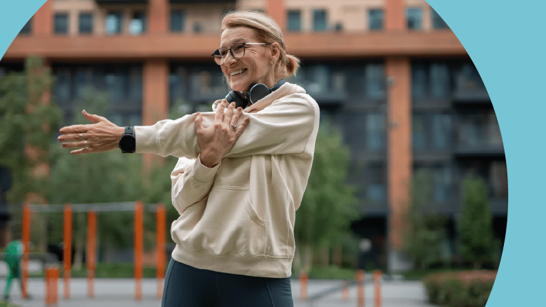 A smiling senior woman warming up her body with an arm stretch in a modern city park.