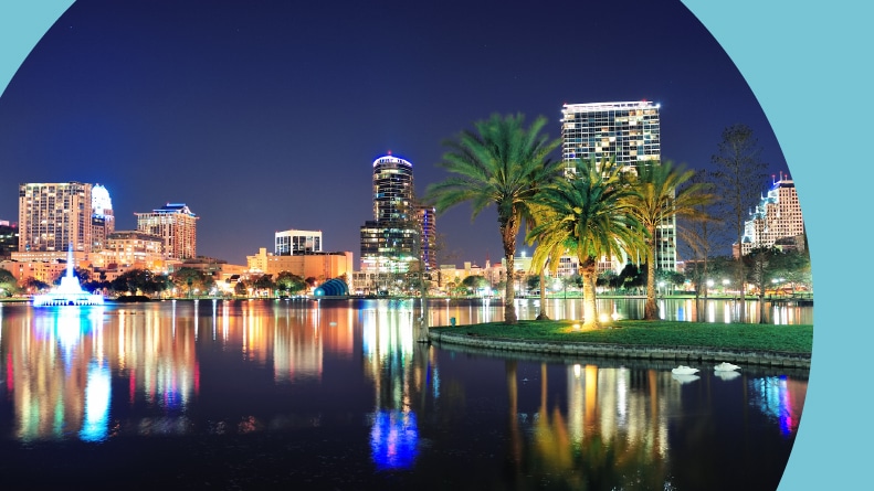View of the Downtown Orlando skyline and Lake Eola at night with skyscrapers, tropic palm trees, and a clear sky.