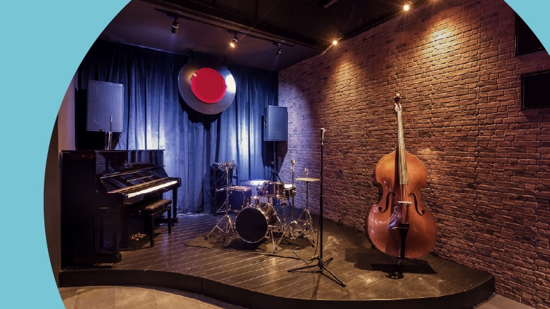 A modern jazz bar with with a black piano, a drum set, and double bass on a stage.