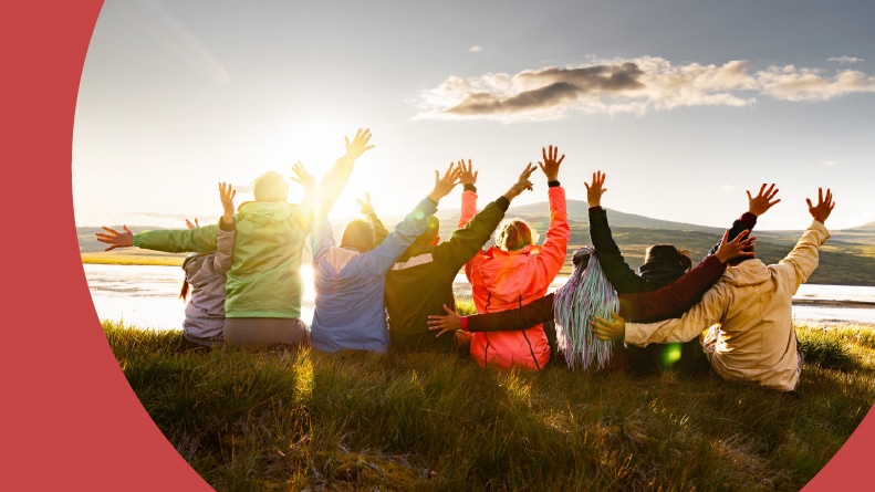 A group of happy 55+ friends sitting by a mountain lake with raised arms at sunset.