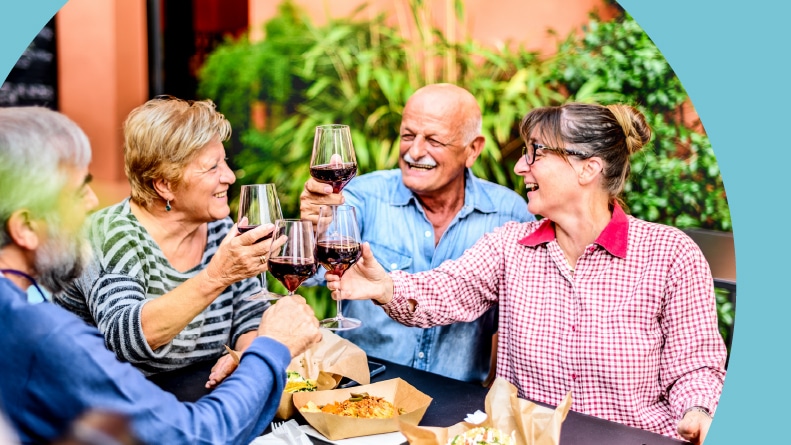 Happy 55+ friends toasting red wine at dinner at a garden party at home.