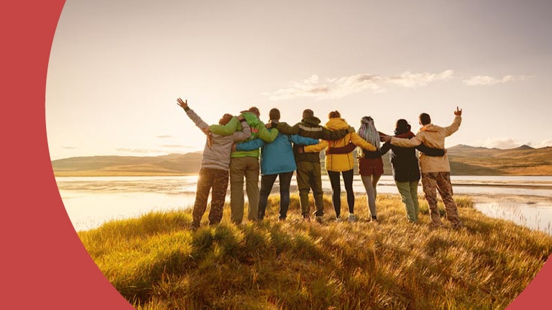 A group of 55+ friends with arms around each other on a hiking trip with a picturesque view.