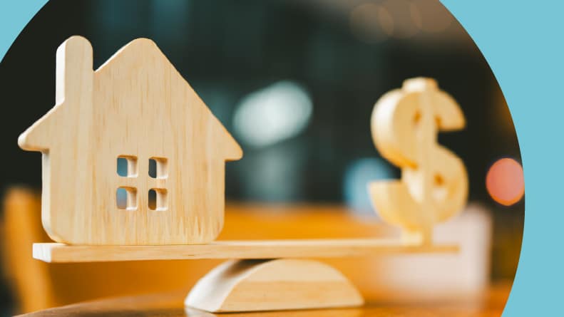 A wood house and dollar sign on a balance scale.