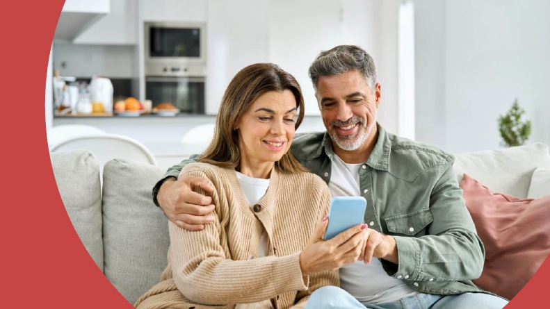 A 55+ couple sitting on a couch looking at a phone.