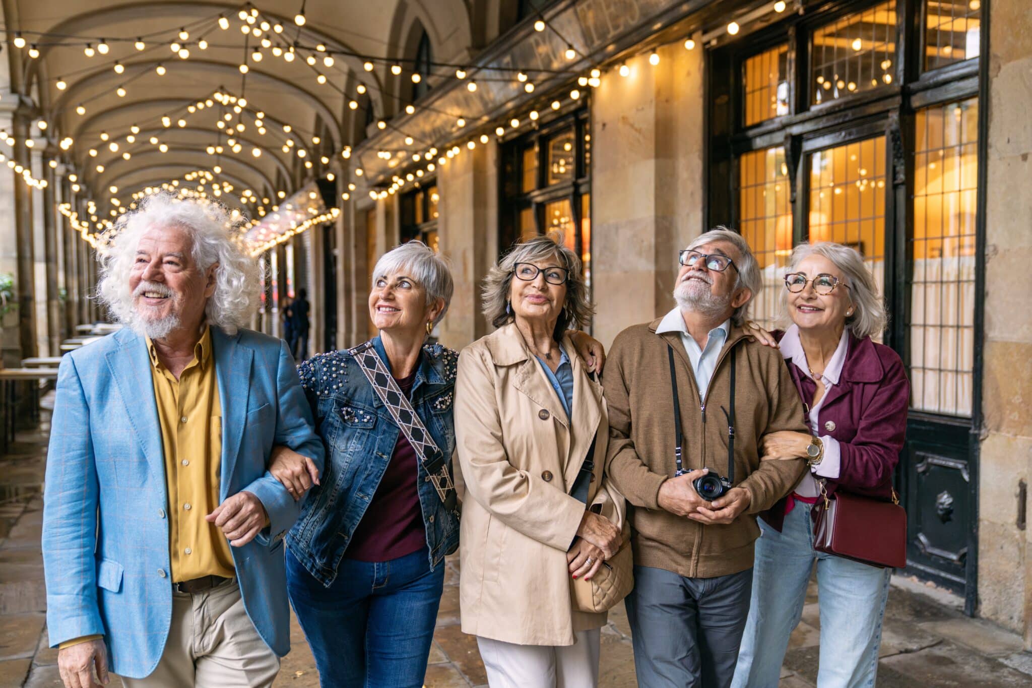 Happy 55+ friends walking arm in arm and exploring a city street lined with festive string lights.