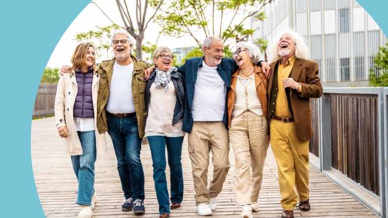 A laughing group of 55+ active adults walking arm in arm down a wooden boardwalk.