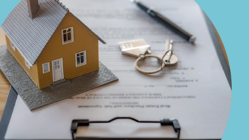 A tiny model of a house on top of a real estate contract beside keys and a pen.