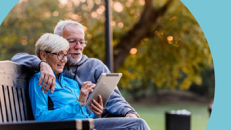 A senior couple sitting on a bench and reading 55+ community reviews on a tablet.