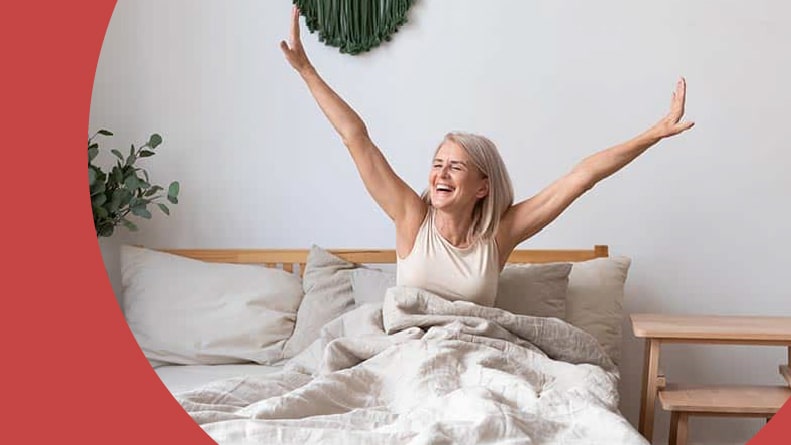 A single 55+ woman stretching and smiling in bed at home.