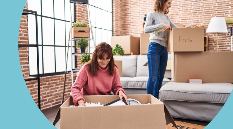 Two women smiling while unpacking cardboard boxes in a bright new home.