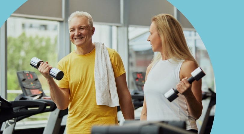 A smiling older man and woman lift dumbbells side by side in a bright, window-lined fitness center at a 55+ community.