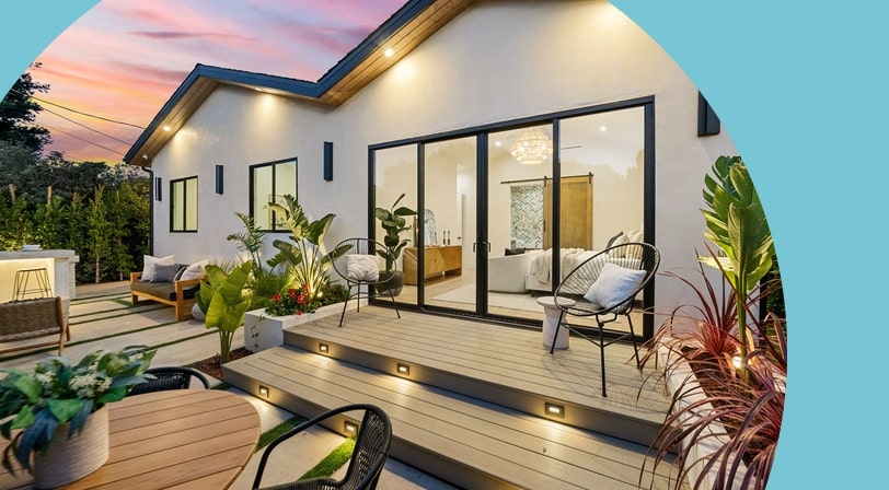 Modern single-story home with a lit wood deck, patio seating, and tropical landscaping at sunset.