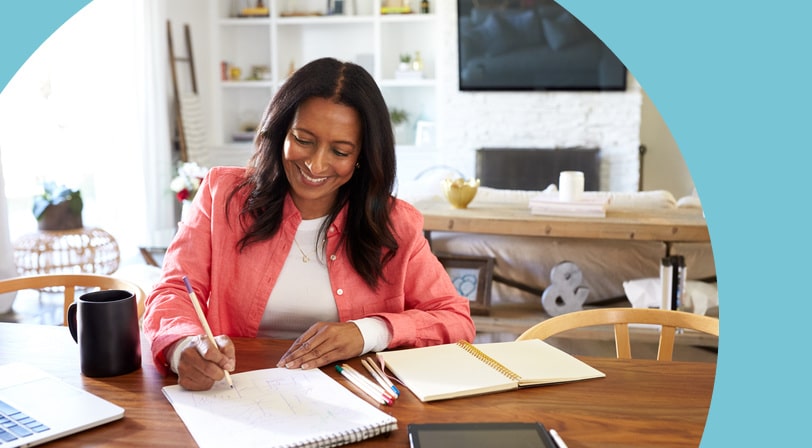 A woman sits at a wooden dining table in a bright, open living space, sketching in a notebook with colored pencils, a laptop and tablet nearby.