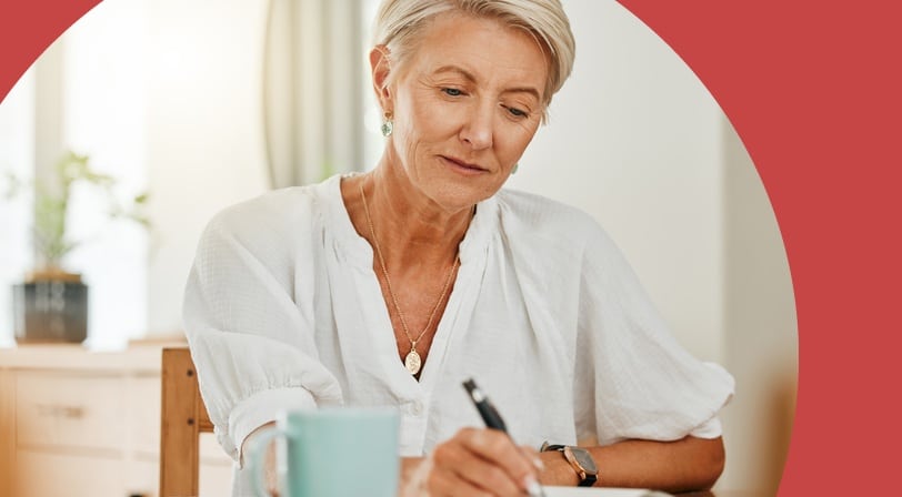 Mature woman writing notes in a notebook at a table with a coffee mug.