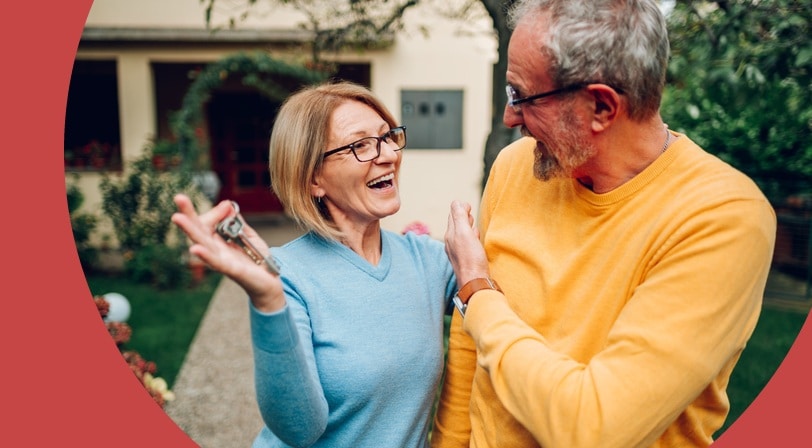 A smiling older couple stand in the front garden of their new home, one of them holding up a set of house keys.
